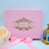 A front-facing view of a rectangular, blush pink velvet invitation box displayed against a clean white background.The lid is centered with a luxurious gold foil crest featuring a crown at the top and intricate floral scrollwork. The name printed in the center of the crest in elegant, clean serif lettering. The box is styled with several decorative elements: A pink satin ribbon tied in a bow at the base of the box. A soft, peach-colored rose partially visible on the left. A small, golden lantern with blue glass panes standing to the right. The combination of the soft velvet texture and shimmering gold foil creates a sophisticated and romantic aesthetic for a special celebration.