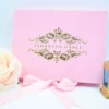 A front-facing view of a rectangular, blush pink velvet invitation box displayed against a clean white background.The lid is centered with a luxurious gold foil crest featuring a crown at the top and intricate floral scrollwork. The name printed in the center of the crest in elegant, clean serif lettering. The box is styled with several decorative elements: A pink satin ribbon tied in a bow at the base of the box. A soft, peach-colored rose partially visible on the left. A small, golden lantern with blue glass panes standing to the right. The combination of the soft velvet texture and shimmering gold foil creates a sophisticated and romantic aesthetic for a special celebration.