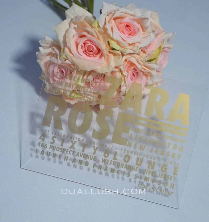features a modern transparent acrylic Bat Mitzvah invitation leaning against a bouquet of soft pink and peach roses. The invitation uses a bold, contemporary design with gold foil text in various sizes. The layout include.