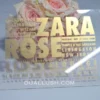 features a modern transparent acrylic Bat Mitzvah invitation leaning against a bouquet of soft pink and peach roses. The invitation uses a bold, contemporary design with gold foil text in various sizes. The layout include.