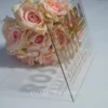 A high-angle, side-view shot of a clear acrylic Bat Mitzvah invitation standing upright on a white surface, positioned in front of a bouquet of peach-colored roses. The invitation features bold, modern typography in a metallic gold or tan color.