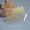 features a modern transparent acrylic Bat Mitzvah invitation leaning against a bouquet of soft pink and peach roses. The invitation uses a bold, contemporary design with gold foil text in various sizes. The layout include.