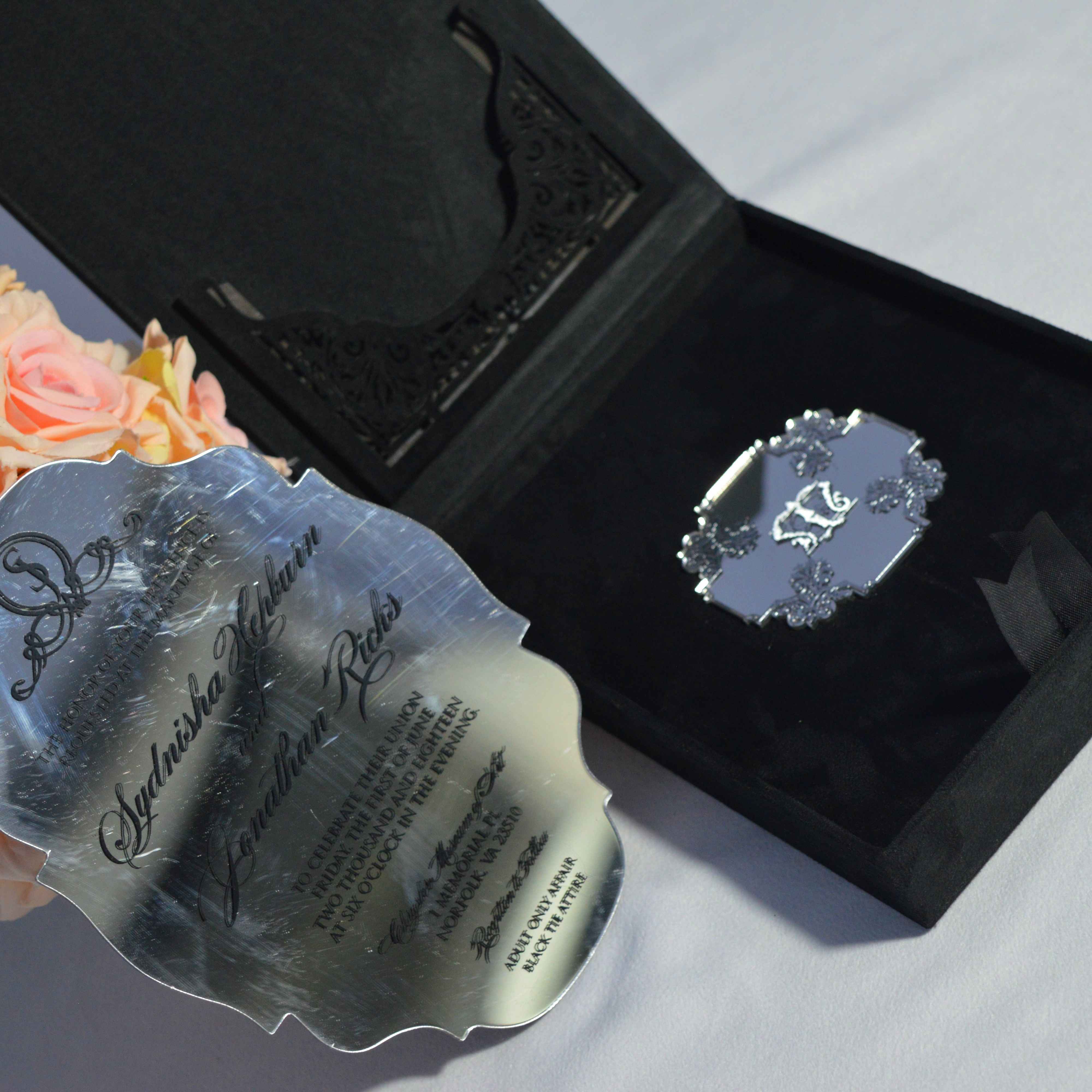 A top-down view of an open champagne silk invitation folio set against a clean white background. The elegant presentation includes:The left panel of the folio featuring a large, ornate silver brooch encrusted with sparkling crystals. A clear acrylic invitation resting in the center, featuring white script and serif typography for the wedding of Decorative accents including a peach-colored rose in the top right and a gold Moroccan-style lantern in the top left.The invitation card details a destination wedding at the Hard Rock Hotel and Casino Punta Cana.