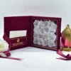 An open burgundy velvet wedding invitation box is shown from a high angle against a white background. The luxurious Sikh wedding suite features: The inside lid: A side-loading pocket on the left panel holds a white enclosure card. The pocket is decorated with a gold foil Khanda symbol** and Gurmukhi script. The base: A recessed compartment on the right holds a clear acrylic invitation card with gold-foiled text. The invitation is uniquely presented over a bed of **white rose petals**, creating a romantic, textured look.Stationery Details: The names are prominently featured in gold serif typography. * **Decorative Accents:** A matching burgundy satin ribbon is draped across the scene. To the right stands a **gold filigree lantern** with pink glass panes, while a white rose is partially visible on the left. The combination of deep burgundy velvet, shimmering gold foil, and fresh white petals creates a regal and culturally rich presentation.