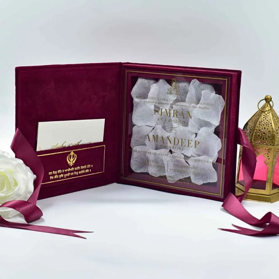 An open burgundy velvet wedding invitation box is shown from a high angle against a white background. The luxurious Sikh wedding suite features: The inside lid: A side-loading pocket on the left panel holds a white enclosure card. The pocket is decorated with a gold foil Khanda symbol** and Gurmukhi script. The base: A recessed compartment on the right holds a clear acrylic invitation card with gold-foiled text. The invitation is uniquely presented over a bed of **white rose petals**, creating a romantic, textured look.Stationery Details: The names are prominently featured in gold serif typography. * **Decorative Accents:** A matching burgundy satin ribbon is draped across the scene. To the right stands a **gold filigree lantern** with pink glass panes, while a white rose is partially visible on the left. The combination of deep burgundy velvet, shimmering gold foil, and fresh white petals creates a regal and culturally rich presentation.