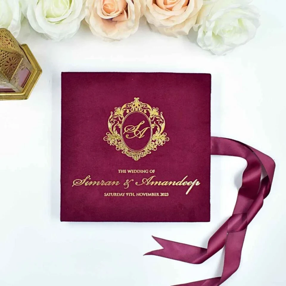 An open maroon velvet wedding invitation box is shown from a high angle against a white background. The luxury suite features: The inside lid: A large, full-color **aerial photograph of a coastal resort** with white buildings, tropical greenery, and bright blue swimming pools bordering the ocean.The base:A recessed compartment holds a **clear acrylic invitation** card. The names are featured in elegant white script, accompanied by formal wedding details. * **Decorative accents:** A matching turquoise fabric band with a **silver wax seal** sits in the base. To the right, a small bronze **Eiffel Tower figurine** stands upright, while a soft **peach rose** is visible on the left. * A silver satin ribbon pull-tab is positioned at the front edge of the box.