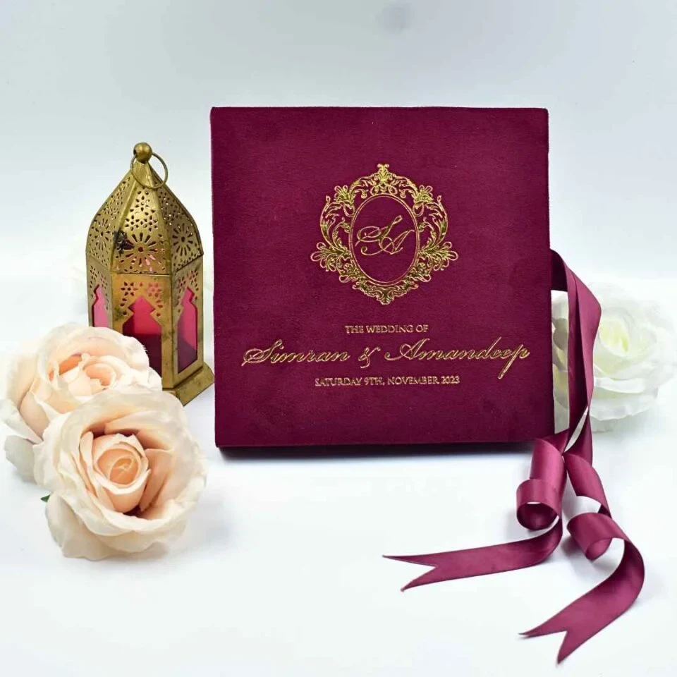 An open maroon velvet wedding invitation box is shown from a high angle against a white background. The luxury suite features: The inside lid: A large, full-color **aerial photograph of a coastal resort** with white buildings, tropical greenery, and bright blue swimming pools bordering the ocean.The base:A recessed compartment holds a **clear acrylic invitation** card. The names are featured in elegant white script, accompanied by formal wedding details. * **Decorative accents:** A matching turquoise fabric band with a **silver wax seal** sits in the base. To the right, a small bronze **Eiffel Tower figurine** stands upright, while a soft **peach rose** is visible on the left. * A silver satin ribbon pull-tab is positioned at the front edge of the box.