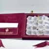 An open burgundy velvet wedding invitation box is shown from a high angle against a white background. The luxurious Sikh wedding suite features: The inside lid: A side-loading pocket on the left panel holds a white enclosure card. The pocket is decorated with a gold foil Khanda symbol** and Gurmukhi script. The base: A recessed compartment on the right holds a clear acrylic invitation card with gold-foiled text. The invitation is uniquely presented over a bed of **white rose petals**, creating a romantic, textured look.Stationery Details: The names are prominently featured in gold serif typography. * **Decorative Accents:** A matching burgundy satin ribbon is draped across the scene. To the right stands a **gold filigree lantern** with pink glass panes, while a white rose is partially visible on the left. The combination of deep burgundy velvet, shimmering gold foil, and fresh white petals creates a regal and culturally rich presentation.