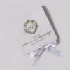 An elegant, square white wedding invitation box or folder, angled diagonally against a light grey background. The top of the box features an intricate gold foil crest with the initials "SA" in a calligraphic script. Below the crest, the text "THE WEDDING OF Simran & Amandeep" is printed in matching gold cursive and serif fonts, followed by the date "SATURDAY 9TH, NOVEMBER 2023." A white satin ribbon is tied into a neat bow at the bottom edge of the box. The lighting creates a soft shadow to the right, emphasizing the texture of the material.