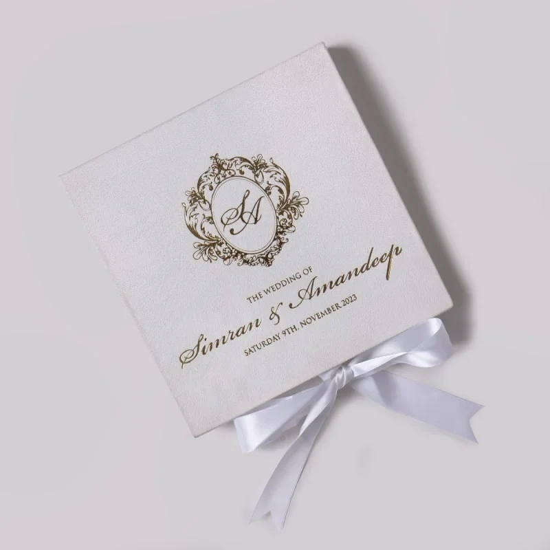 An elegant, square white wedding invitation box or folder, angled diagonally against a light grey background. The top of the box features an intricate gold foil crest with the initials "SA" in a calligraphic script. Below the crest, the text "THE WEDDING OF Simran & Amandeep" is printed in matching gold cursive and serif fonts, followed by the date "SATURDAY 9TH, NOVEMBER 2023." A white satin ribbon is tied into a neat bow at the bottom edge of the box. The lighting creates a soft shadow to the right, emphasizing the texture of the material.