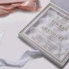 An open, square wedding invitation box in white with gold accents. The main invitation is printed on a clear acrylic or vellum overlay, resting on a bed of delicate white silk flower petals. The text, in gold foil, announces the wedding of Simran and Amandeep, featuring their names in a large serif font and details of the event below. The inner lid of the box contains a pocket holding additional cards, including an RSVP and accommodation details. Beside the box, a portion of a blush pink satin ribbon and another card with a decorative crest are visible. The entire suite is set against a clean white background, creating a luxurious and airy aesthetic.