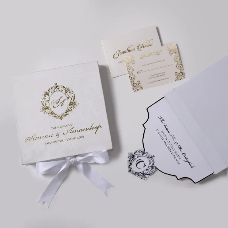 An elegant, square white wedding invitation box or folder, angled diagonally against a light grey background. The top of the box features an intricate gold foil crest with the initials "SA" in a calligraphic script. Below the crest, the text "THE WEDDING OF Simran & Amandeep" is printed in matching gold cursive and serif fonts, followed by the date "SATURDAY 9TH, NOVEMBER 2023." A white satin ribbon is tied into a neat bow at the bottom edge of the box. The lighting creates a soft shadow to the right, emphasizing the texture of the material.