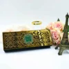 A long, rectangular emerald green Scroll invitation box featuring a highly reflective gold laser-cut overlay with intricate heart and scroll patterns. In the center of the gold plate, a small green square features an embossed silver monogram. The box is set on a white surface, accompanied by a small bronze Eiffel Tower figurine, a cluster of peach roses, and a gold-rimmed glass. The bright lighting highlights the mirror-like finish of the gold decorative panel.