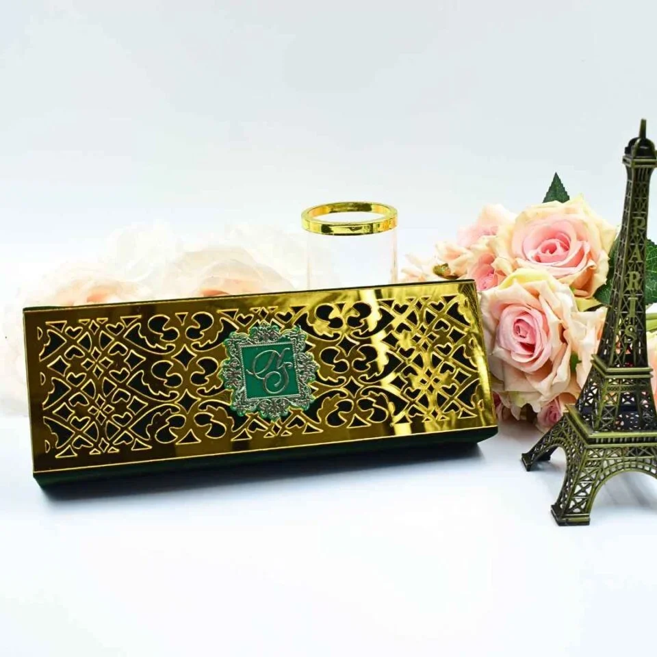 A long, rectangular emerald green Scroll invitation box featuring a highly reflective gold laser-cut overlay with intricate heart and scroll patterns. In the center of the gold plate, a small green square features an embossed silver monogram. The box is set on a white surface, accompanied by a small bronze Eiffel Tower figurine, a cluster of peach roses, and a gold-rimmed glass. The bright lighting highlights the mirror-like finish of the gold decorative panel.