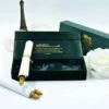 A high-angle, close-up shot of an elegant emerald green and gold wedding invitation suite. The main focus is an unrolled **green fabric scroll** featuring gold calligraphy that reads "Naveed & Sahar" and "Mehndi Ceremony." The text indicates the event is on Thursday, December 21, 2023. The scroll is displayed alongside: * A matching **emerald green velvet box** with a gold prayer inscribed on the inner lid. * A second closed box featuring a gold "NS" monogram. * Two white roses and a small bronze Eiffel Tower figurine in the background. * A white paper scroll with gold finials resting in the foreground. The entire arrangement is set against a clean, white background, emphasizing the luxurious jewel tones and metallic accents.
