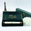 A high-angle, close-up shot of an elegant emerald green and gold wedding invitation suite. The main focus is an unrolled **green fabric scroll** featuring gold calligraphy that reads "Naveed & Sahar" and "Mehndi Ceremony." The text indicates the event is on Thursday, December 21, 2023. The scroll is displayed alongside: * A matching **emerald green velvet box** with a gold prayer inscribed on the inner lid. * A second closed box featuring a gold "NS" monogram. * Two white roses and a small bronze Eiffel Tower figurine in the background. * A white paper scroll with gold finials resting in the foreground. The entire arrangement is set against a clean, white background, emphasizing the luxurious jewel tones and metallic accents.