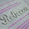 A close-up, angled shot of a sparkling silver glitter Bat Mitzvah invitation. The name is featured prominently in large, raised silver script. The surrounding text is printed in a vibrant magenta font, framed by a thin magenta border. In the blurred background, a bouquet of peach and pink roses is visible.