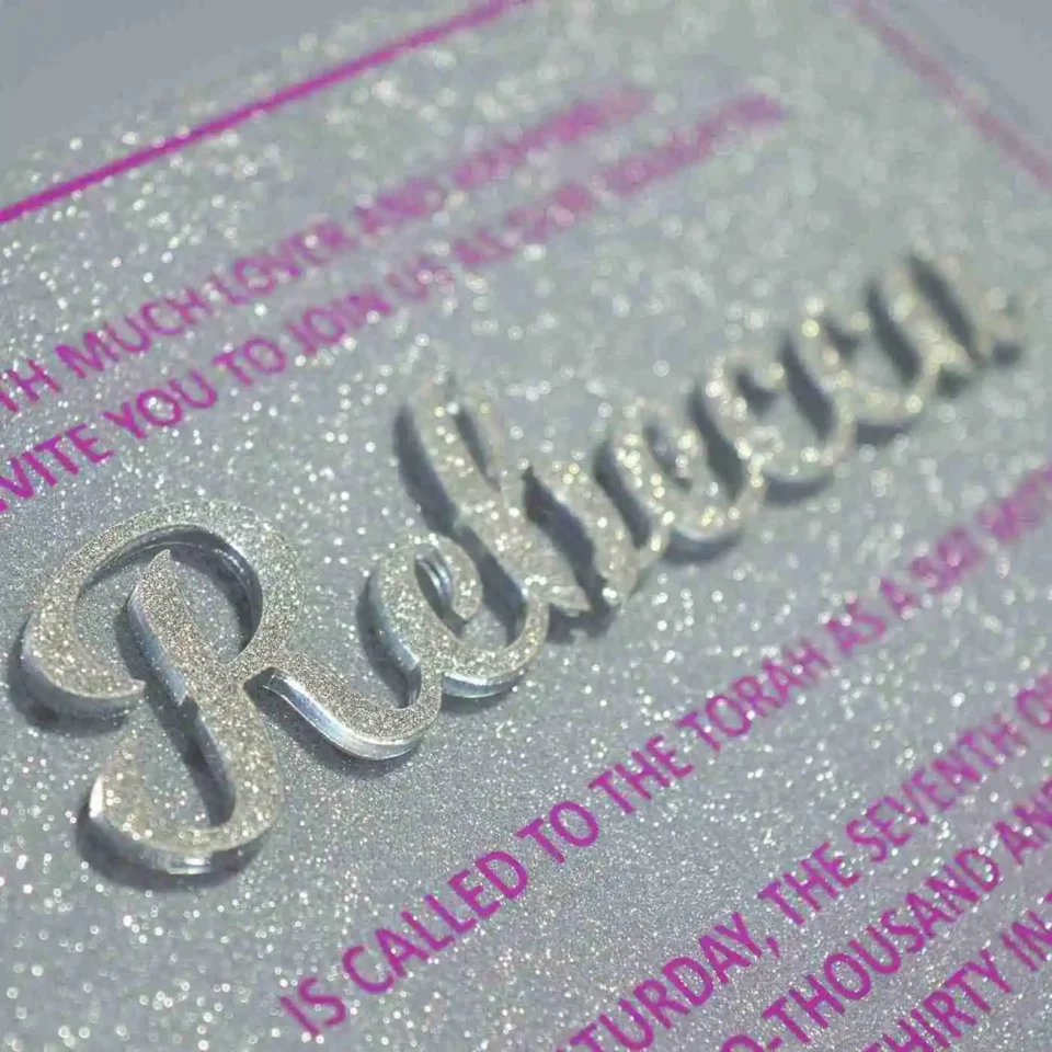 A close-up, angled shot of a sparkling silver glitter Bat Mitzvah invitation. The name is featured prominently in large, raised silver script. The surrounding text is printed in a vibrant magenta font, framed by a thin magenta border. In the blurred background, a bouquet of peach and pink roses is visible.