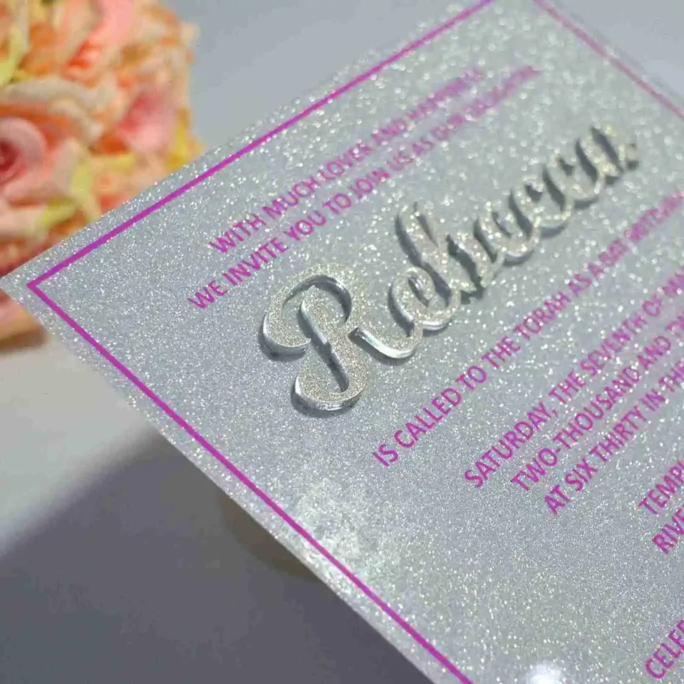 A close-up, angled shot of a sparkling silver glitter Bat Mitzvah invitation. The name is featured prominently in large, raised silver script. The surrounding text is printed in a vibrant magenta font, framed by a thin magenta border. In the blurred background, a bouquet of peach and pink roses is visible.