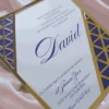 A white anAXd blue acrylic Bar Mitzvah invitation with an intricate gold laser-cut Art Deco border is displayed on a shimmering dusty rose silk fabric. A small mustard navy blue envelope sits behind it. The invitation features a Star of David at the top and the following details in blue script and serif fonts.