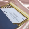 A luxurious Bar Mitzvah invitation partially tucked into a navy blue envelope. The invitation features a white card with navy blue text for "Jonathan," bordered by a thick, polished gold acrylic frame. The top corner of the frame boasts an intricate, laser-cut geometric lattice design. The ensemble rests on a shimmering, draped champagne pink satin fabric.