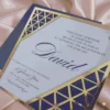 A white and blue acrylic Bar Mitzvah invitation with an intricate gold laser-cut Art Deco border is displayed on a shimmering dusty rose silk fabric. A small mustard navy blue envelope sits behind it. The invitation features a Star of David at the top and the following details in blue script and serif fonts.