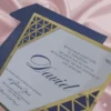 A white and blue acrylic Bar Mitzvah invitation with an intricate gold laser-cut Art Deco border is displayed on a shimmering dusty rose silk fabric. A small mustard navy blue envelope sits behind it. The invitation features a Star of David at the top and the following details in blue script and serif fonts.