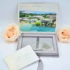 An open, **luxurious destination wedding invitation box** in soft grey velvet. The suite features: * **Inner lid artwork:** A vibrant, full-color aerial photograph of a tropical beach resort with turquoise water and palm trees. * **Acrylic invitation:** A clear acrylic card with white calligraphy for "Kirsten & Danielle" rests in the velvet-lined base, adorned with a decorative silver-grey silk tassel. * **Supporting stationery:** A cream-colored envelope in the foreground addressed to "Carmella & Arthur" in silver foil script. * **Accents:** Two large, pale peach roses frame the box on either side against a clean white background. * A matching grey silk ribbon pull-tab is visible at the bottom of the velvet box.