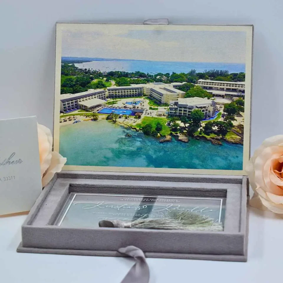 An open, **luxurious destination wedding invitation box** in soft grey velvet. The suite features: * **Inner lid artwork:** A vibrant, full-color aerial photograph of a tropical beach resort with turquoise water and palm trees. * **Acrylic invitation:** A clear acrylic card with white calligraphy for "Kirsten & Danielle" rests in the velvet-lined base, adorned with a decorative silver-grey silk tassel. * **Supporting stationery:** A cream-colored envelope in the foreground addressed to "Carmella & Arthur" in silver foil script. * **Accents:** Two large, pale peach roses frame the box on either side against a clean white background. * A matching grey silk ribbon pull-tab is visible at the bottom of the velvet box.