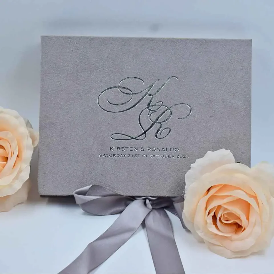 A rectangular, grey suede-textured wedding invitation box sits centered on a white surface. The front of the box features a large, elegant silver foil monogram of the initials "K & R" in interlocking script, with the names "KIRSTEN & RONALDO" and the date "SATURDAY 21ST OF OCTOBER 2023" printed in smaller silver serif font below. A matching grey satin ribbon is tied into a bow at the bottom. The box is flanked by two large, pale peach roses, and the background is a clean, cool white.