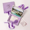 An open, **luxurious lavender velvet invitation box** designed for a destination wedding, shown against a bright white background. The interior of the box features: * **Inner lid artwork:** A stunning, full-color aerial photograph of a tropical beach resort with bright turquoise water and palm trees, framed by silver ornate corners. * **Acrylic invitation:** A clear acrylic card with elegant script calligraphy sits in the velvet-lined base, accented by a **long, purple silk tassel** hanging from the center. * **Supporting stationery:** A cream-colored card with silver foil text is visible behind the clear acrylic. * **Decorative accents:** A large **pale peach rose** on the left and a small **bronze Eiffel Tower figurine** on the right frame the presentation. * A matching purple silk ribbon pull-tab is visible at the top of the box.