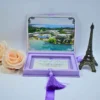 An open, **luxurious lavender velvet invitation box** designed for a destination wedding, shown against a bright white background. The interior of the box features: * **Inner lid artwork:** A stunning, full-color aerial photograph of a tropical beach resort with bright turquoise water and palm trees, framed by silver ornate corners. * **Acrylic invitation:** A clear acrylic card with elegant script calligraphy sits in the velvet-lined base, accented by a **long, purple silk tassel** hanging from the center. * **Supporting stationery:** A cream-colored card with silver foil text is visible behind the clear acrylic. * **Decorative accents:** A large **pale peach rose** on the left and a small **bronze Eiffel Tower figurine** on the right frame the presentation. * A matching purple silk ribbon pull-tab is visible at the top of the box.