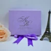 A front-facing, centered shot of a **lavender textured fabric invitation box** featuring silver foil details. The box is designed with: * A large, elegant **silver foil "KR" monogram** at the center. * The names "Kirsten & Ronaldo" and the wedding date "Saturday 21st of October 2023" printed in silver foil below the monogram. * A bright **purple satin ribbon** tied in a bow at the bottom of the box. * The box is flanked by a large **pale peach rose** on the left and a small **bronze Eiffel Tower figurine** on the right. * The entire arrangement is set against a clean, white background, highlighting the soft lavender tones and shimmering silver accents.
