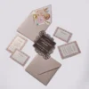 An elegant Quinceañera invitation suite featuring a dusty rose and rose gold color palette, laid out on a light gray background. The set includes several coordinated pieces: Main Invitation: A square, chocolate-brown card with a decorative rose gold laser-cut floral border made of rose patterns. The white and rose gold text reads, "PLEASE JOIN US FOR A QUINCEAÑERA celebration IN HONOR OF ASHLEY JOHNSON" with the date "11.24.2024." Enclosure Cards: Three white cards featuring a matching rose gold floral pattern around the edges. These include two "Response" (RSVP) cards and one "Details" card for accommodations. Envelope: A dusty rose envelope is shown open, revealing a luxurious floral liner featuring large painted roses in shades of cream, pink, and gold with gold leaf accents. A second invitation card is partially tucked inside.