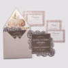 An elegant Quinceañera invitation suite featuring a dusty rose and rose gold color palette, laid out on a light gray background. The set includes several coordinated pieces: Main Invitation: A square, chocolate-brown card with a decorative rose gold laser-cut floral border made of rose patterns. The white and rose gold text reads, "PLEASE JOIN US FOR A QUINCEAÑERA celebration IN HONOR OF ASHLEY JOHNSON" with the date "11.24.2024." Enclosure Cards: Three white cards featuring a matching rose gold floral pattern around the edges. These include two "Response" (RSVP) cards and one "Details" card for accommodations. Envelope: A dusty rose envelope is shown open, revealing a luxurious floral liner featuring large painted roses in shades of cream, pink, and gold with gold leaf accents. A second invitation card is partially tucked inside.