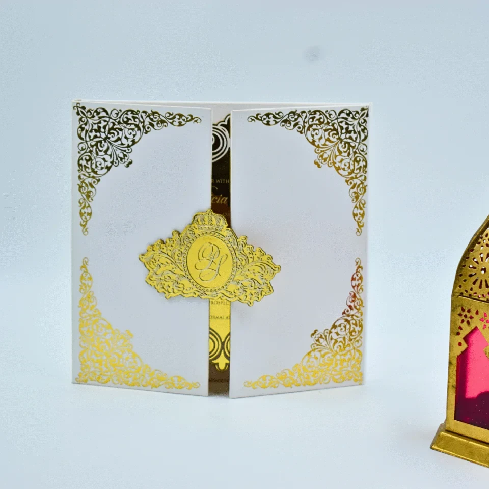 An elegant white gatefold wedding invitation standing upright on a white surface. The invitation features intricate gold foil filigree scrollwork decorating all four corners of the front panels. The two panels are joined by a central, die-cut gold crown seal featuring a monogrammed. The panels are slightly open, revealing a glimpse of a black and gold invitation card inside. To the right, a gold Moroccan-style lantern with magenta glass panels adds a decorative accent to the scene.