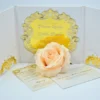 An elegant wedding invitation suite set against a clean white background. The central piece is a white tri-fold folder featuring an ornate, laser-cut gold acrylic frame in the center. Inside the gold frame, the invitation text is engraved, naming the couple as.