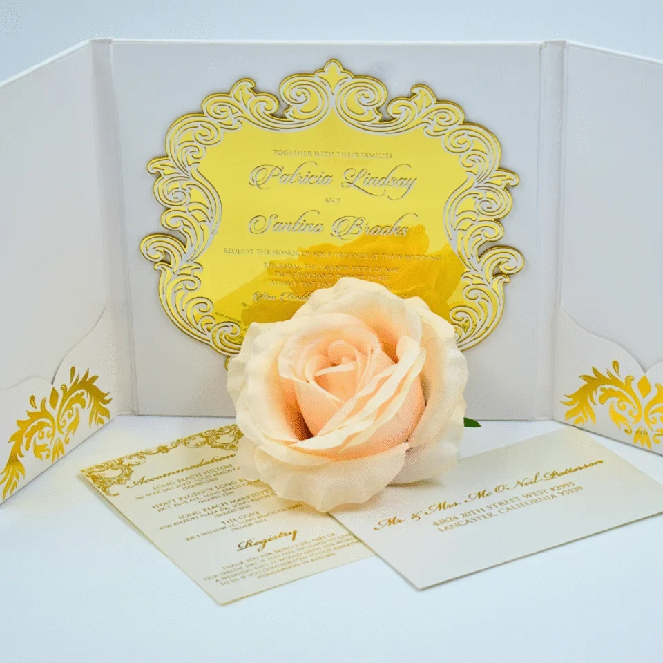 An elegant wedding invitation suite set against a clean white background. The central piece is a white tri-fold folder featuring an ornate, laser-cut gold acrylic frame in the center. Inside the gold frame, the invitation text is engraved, naming the couple as.
