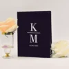 An elegant, black suede or velvet folio-style invitation stands upright against a plain white background. The front of the folio is personalized with a white or silver monogram featuring a large serif letter separated by a horizontal line. Below the monogram, the date is printed in a clean, smaller font.