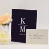 A sophisticated wedding invitation suite featuring a **black velvet folio** with a large, white serif monogram separated by a horizontal line, with the date printed below. Resting in front is a textured cream envelope addressed to in elegant black script. To the left, a soft peach rose in a glass vase and a white rose add a delicate, romantic touch to the monochromatic design.