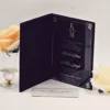 An open, black velvet folio wedding invitation acrylic set against a white background. The right side features a sleek, black acrylic invitation card with acrylic silver engraved text and a monogram at the top. The names written in an elegant script. The wedding details list the date as print. The left side of the folio has a pocket containing a white RSVP or detail card with the names visible in script. The invitation is accessorized with a peach-colored rose in a small gold-rimmed glass pedestal vase to the left and a white rose lying on the surface to the right.An open, black velvet folio wedding invitation acrylic set against a white background. The right side features a sleek, black acrylic invitation card with acrylic silver engraved text and a monogram at the top. The names written in an elegant script. The wedding details list the date as print. The left side of the folio has a pocket containing a white RSVP or detail card with the names visible in script. The invitation is accessorized with a peach-colored rose in a small gold-rimmed glass pedestal vase to the left and a white rose lying on the surface to the right.