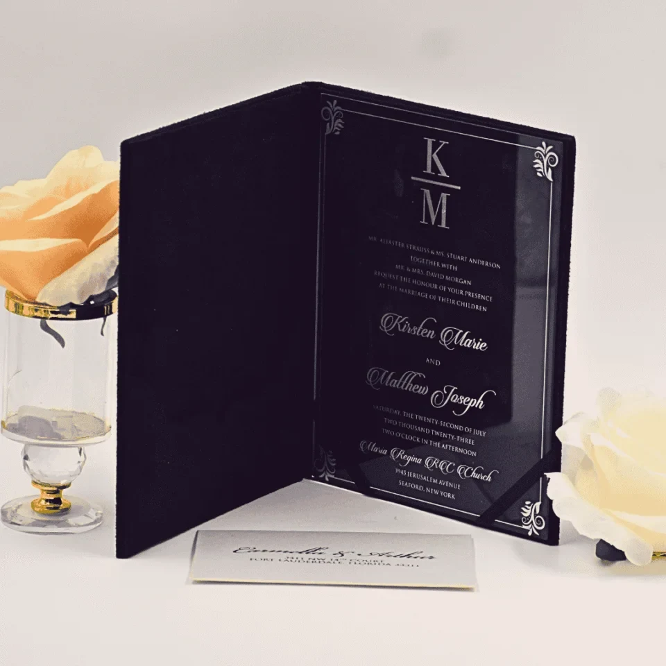 An open, black velvet folio wedding invitation acrylic set against a white background. The right side features a sleek, black acrylic invitation card with acrylic silver engraved text and a monogram at the top. The names written in an elegant script. The wedding details list the date as print. The left side of the folio has a pocket containing a white RSVP or detail card with the names visible in script. The invitation is accessorized with a peach-colored rose in a small gold-rimmed glass pedestal vase to the left and a white rose lying on the surface to the right.An open, black velvet folio wedding invitation acrylic set against a white background. The right side features a sleek, black acrylic invitation card with acrylic silver engraved text and a monogram at the top. The names written in an elegant script. The wedding details list the date as print. The left side of the folio has a pocket containing a white RSVP or detail card with the names visible in script. The invitation is accessorized with a peach-colored rose in a small gold-rimmed glass pedestal vase to the left and a white rose lying on the surface to the right.