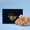 A luxurious black velvet gatefold invitation suite displayed against a light blue background. The folio is partially open, revealing a striking gold mirror acrylic invitation inside with black engraved text. A large, ornate gold mirror acrylic monogram, featuring the initials in a swirling script, is attached to the left flap of the velvet folio.