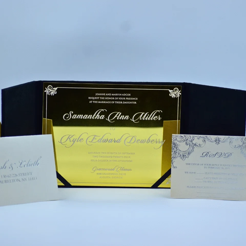 A full wedding invitation suite featuring a central **gold mirror acrylic invitation held within an open **black velvet gatefold folio**. The card is printed with white script and delicate corner flourishes. In the foreground, a cream-colored envelope addressed to **"Josh & Lelieth"** sits on the left, while a matching **RSVP card** with ornate navy blue scrollwork is positioned on the right. The entire set is displayed against a clean, light blue background.