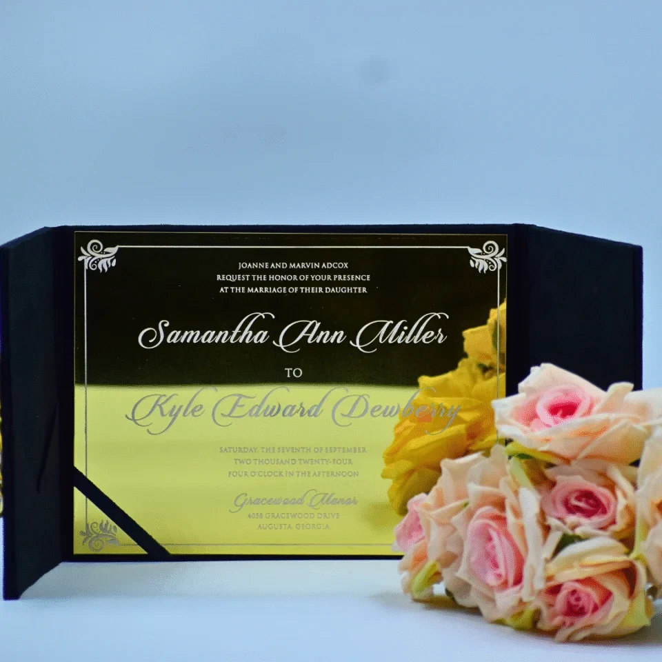 An elegant wedding invitation for displayed within a **black velvet gatefold folio**. The invitation is printed on a highly reflective **gold mirror acrylic** card, featuring white script and delicate silver corner flourishes. To the right of the invitation, a lush bouquet of **peach and pink roses** adds a soft, romantic contrast to the bold, metallic stationery. The set is photographed against a clean, light blue background.