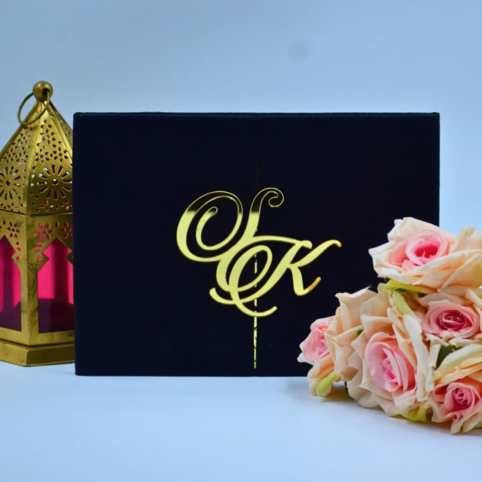 A luxurious black velvet gatefold invitation suite displayed against a light blue background. The folio is partially open, revealing a striking gold mirror acrylic invitation inside with black engraved text. A large, ornate gold mirror acrylic monogram, featuring the initials in a swirling script, is attached to the left flap of the velvet folio.