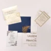A sophisticated wedding invitation suite centered around a transparent acrylic invitation. The main card is partially tucked into a navy blue envelope with a shimmering metallic gold liner.