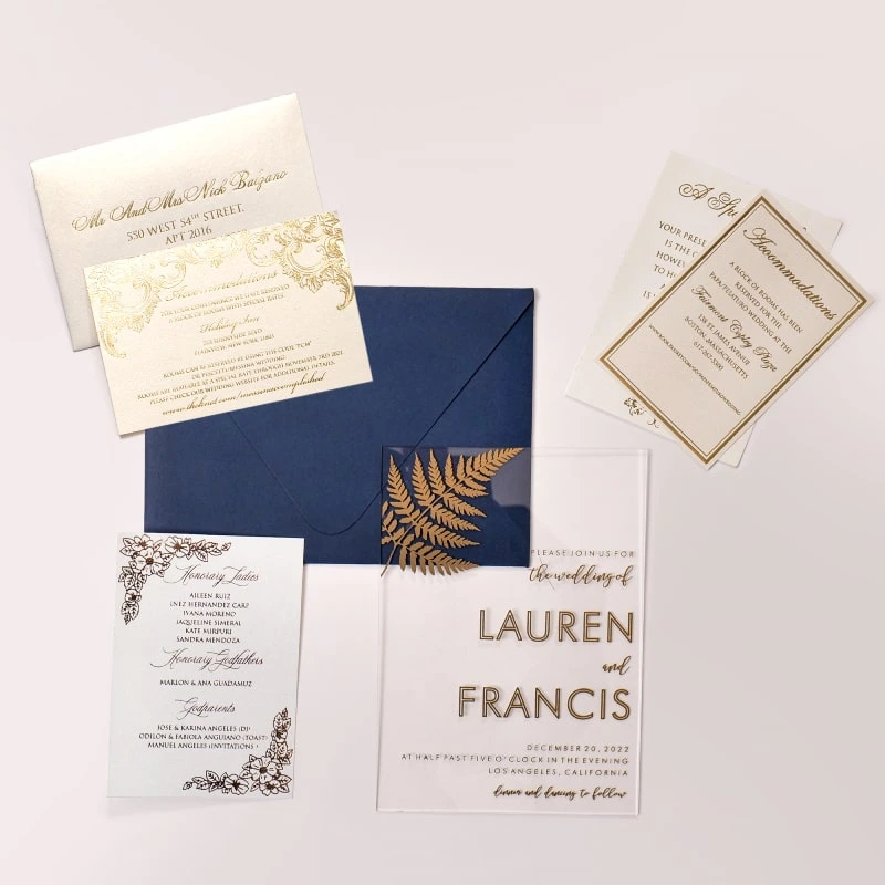 A sophisticated wedding invitation suite centered around a transparent acrylic invitation. The main card is partially tucked into a navy blue envelope with a shimmering metallic gold liner.