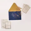 A sophisticated wedding invitation suite centered around a transparent acrylic invitation. The main card is partially tucked into a navy blue envelope with a shimmering metallic gold liner.