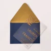 A sophisticated wedding invitation suite centered around a transparent acrylic invitation. The main card is partially tucked into a navy blue envelope with a shimmering metallic gold liner.